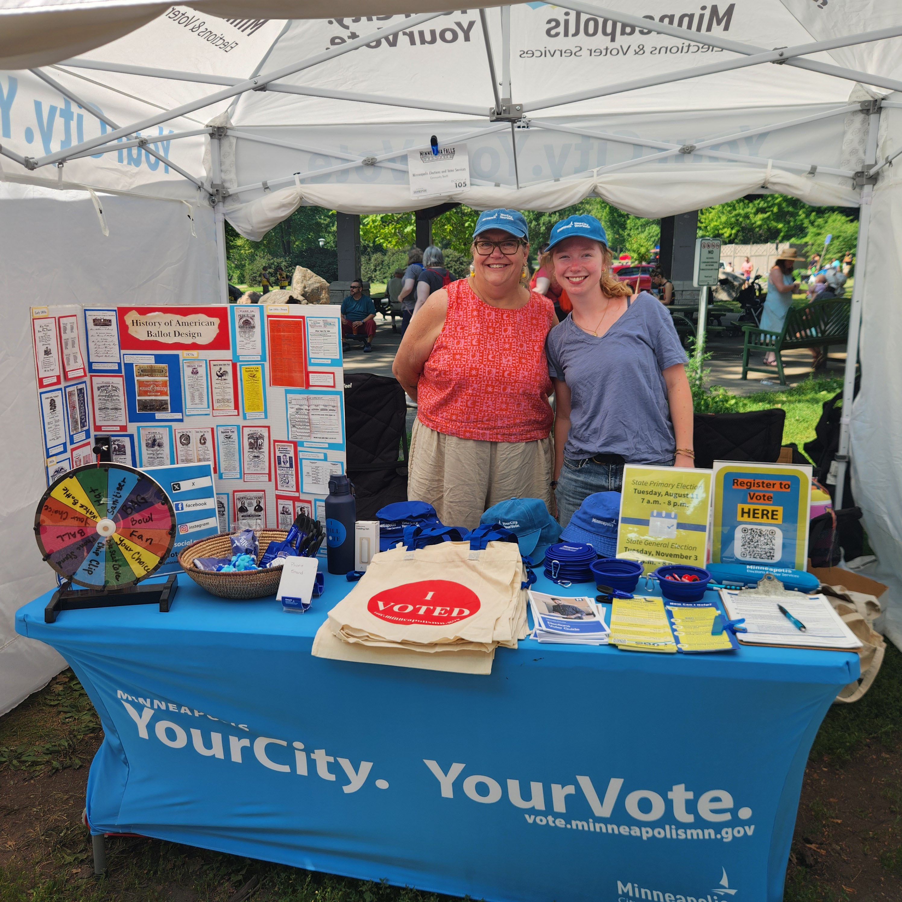 Two Minneapolis Elections & Voter Services staff smile together in front of a table at an art fair. The table includes a poster showcasing the history of American ballot design, a spinning wheel with prizes around it, Voter Registration Applications, and flyers with information about elections.